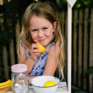 Ein kleines Mädchen steht im Garten eines Kindergartens hinter einem selbstgemachten Limonadenstand. Vor ihr auf dem Tisch stehen eine Schale mit einer Zitrone und ein Becher. In der Hand hält sie eine angebissene Zitrone und verzieht das Gesicht zu einer witzigen Mischung aus Grimasse und Lächeln, während die Szene sommerlich und lebendig wirkt.